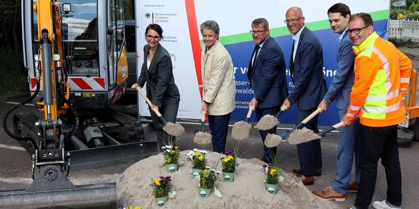Sechs Personen schippen Sand vor einem Bagger.