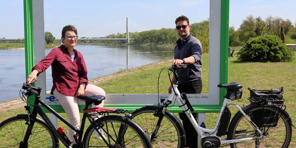 Im Rahmen der Aktion Stadtradeln werden mehrere Touren angeboten. © Stadt Schönebeck (Elbe) Zwei Menschen stehen an einem großen Bilderrahmen vor der Elbe und der Elbauenbrücke.