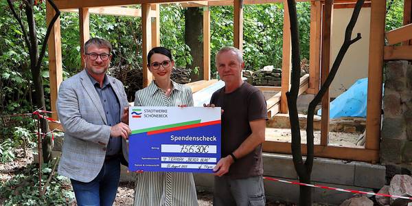 Julia Teige, Geschäftsführerin der Stadtwerke Schönebeck GmbH, übergab einen Spendenscheck an Zootierpfleger Uwe Schlöcker (r.) und Oberbürgermeister Bert Knoblauch. © Stadt Schönebeck (Elbe) Ein großer symbolsicher Scheck wird von einer Frau und zwei Männern vor der Baustelle für ein neues Kleintierhaus festgehalten.
