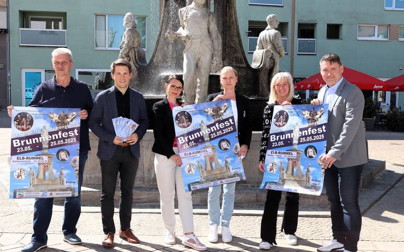 Sechs personen stehen vor einem Brunnen und präsentieren Plakate für die Veranstaltung.