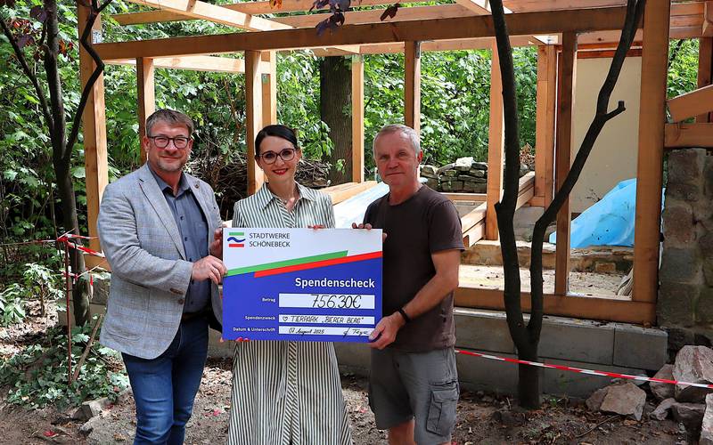 Ein großer symbolsicher Scheck wird von einer Frau und zwei Männern vor der Baustelle für ein neues Kleintierhaus festgehalten.